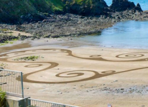 A beach scene with intricate spiral and wave patterns drawn in the sand, reflecting the unique sand art styles of Copper Coast. The designs are surrounded by rocks and water, with a grassy hillside and a railing in the foreground.