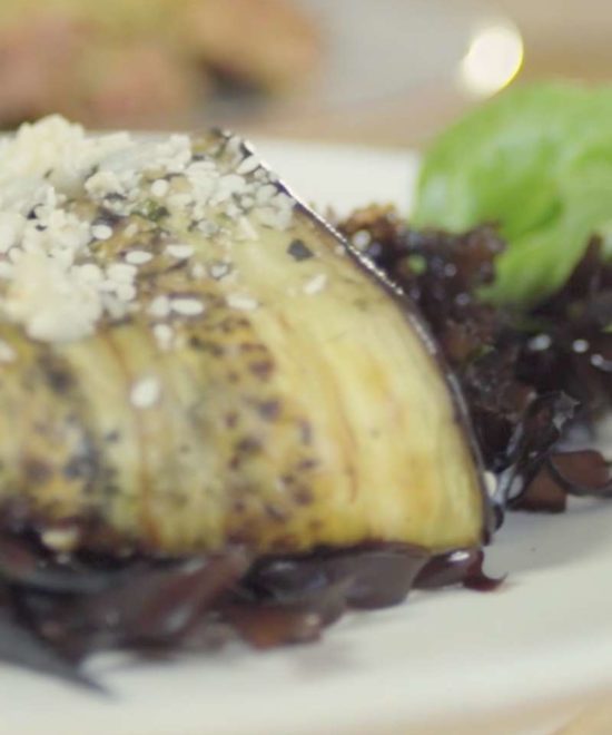 A close-up of a stuffed eggplant slice, adorned with crumbled cheese and served on a bed of dark greens, conjures images of an adventurous day spent seaweed foraging along the Copper Coast. A fresh basil leaf peeks out in the background.