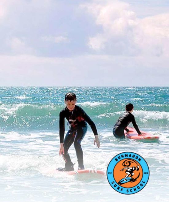 Two young surfers in wetsuits ride small waves on bright red surfboards along the stunning Copper Coast. The sunny sea sparkles under a blue sky with scattered clouds, as the Bunmahon Surf School logo is visible in the corner.