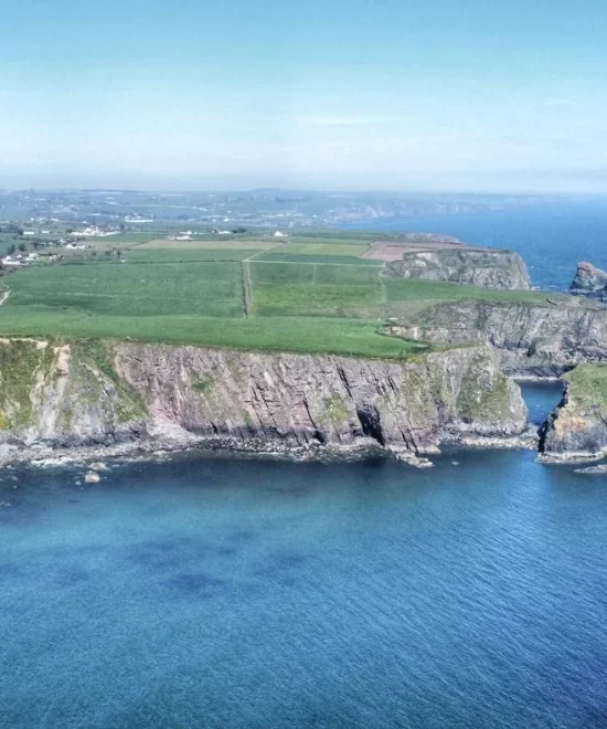 Aerial view of Ballydwan Bay on the Copper Coast, showcasing a coastal landscape with cliffs and caves beside a calm, blue sea. Green fields stretch back from the cliff edge under a clear sky, while small rocky islands dot the water near the shore.