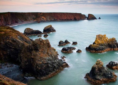 A scenic view of Ballydwan Bay on the Copper Coast features large, jagged rock formations in the foreground, surrounded by calm sea waters. The background showcases a dramatic cliff under a cloudy sky, creating a serene and picturesque landscape.