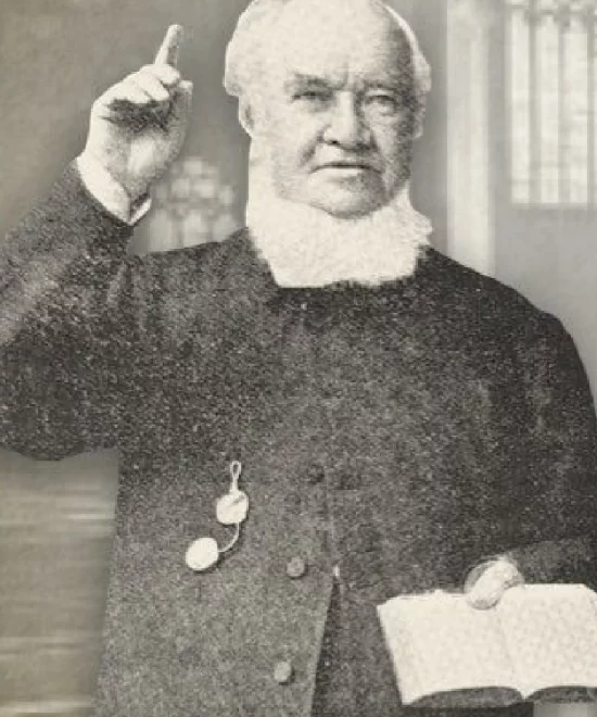 A black and white photo features David Alfred Doudney, an older man with white hair and sideburns, clad in a dark suit. He holds an open book and raises one finger, standing against a blurred indoor backdrop reminiscent of Copper Coast's understated elegance.