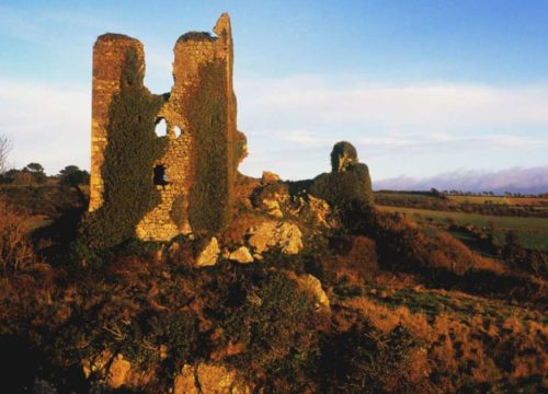 A ruined stone tower, reminiscent of Dunhill Castle, stands on a rocky hill covered in ivy and bathed in warm sunset light. The landscape along the Copper Coast features grassy fields and distant trees beneath a partly cloudy sky.