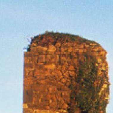 A tall, rocky cliff with a flat top, partially covered in greenery, stands against a clear blue sky along the Copper Coast. The warm light gives the rock a golden hue.