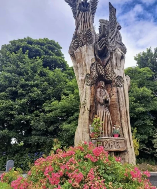 A large, intricate wooden sculpture stands in front of lush green trees and vibrant pink flowers in Fenor village. The carving, embodying the rich history of Fenor village, features several figures, including a central human figure and birds above. The word "HOPE" is carved at the base.