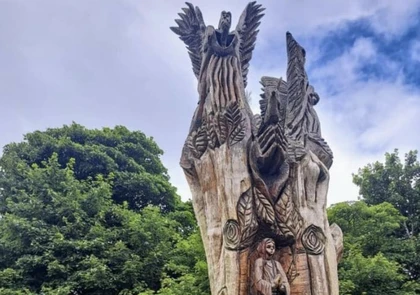 A large wooden sculpture with detailed carvings of leaves, a person, and winged figures stands majestically, echoing the rich history of Fenor village. It is set against a backdrop of lush green trees and a partly cloudy sky.