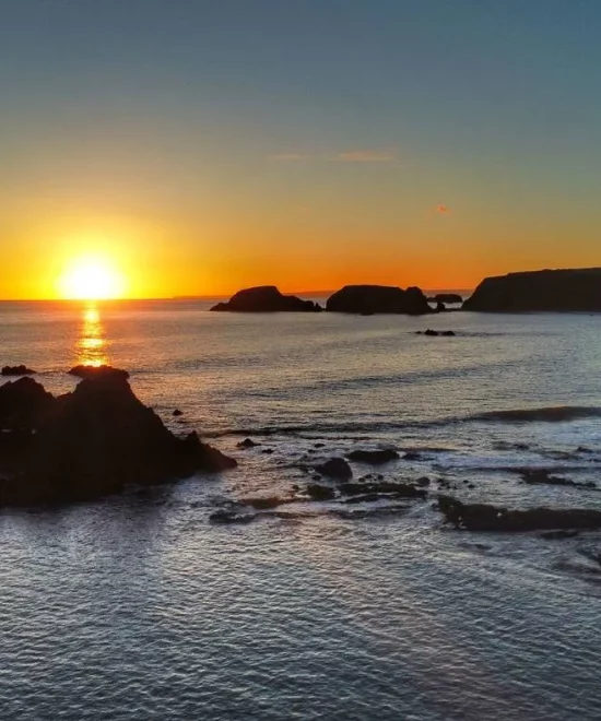 A sunset over Garrarus Strand on the Copper Coast graces the clear sky. The sun nears the horizon, casting a warm glow. Rocky formations and cliffs are silhouetted against the vibrant orange and blue hues, reflecting off the calm sea.