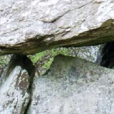 A close-up of large, weathered rocks stacked together, reminiscent of the Gaulstown Dolmen, forming a small archway. The rocks are rough and have a mix of gray and brown tones, with patches of moss. Background shows a hint of greenery typical of the Copper Coast.