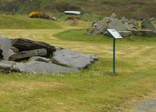 A grassy landscape in the Geological Garden of Copper Coast features large rocks arranged in a circular formation, with an informational sign nearby. Rolling hills are visible in the background, adding to the area's natural beauty.