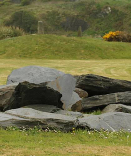 A pile of large, gray stones sits majestically on a grassy field in the Geological Garden. The scene is beautifully framed by green hills and patches of yellow flowers, echoing the natural splendor of Copper Coast.