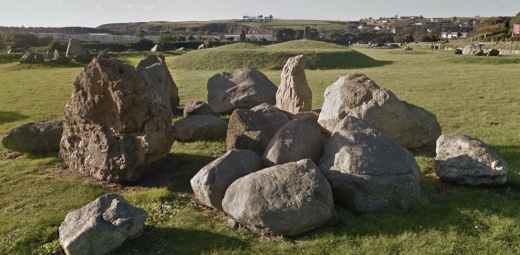 A circle of large, weathered boulders is artfully arranged in a grassy field under the brooding sky of Geological Garden. In the distance, the rolling hills of Copper Coast and distant buildings enhance the scene's rugged beauty.