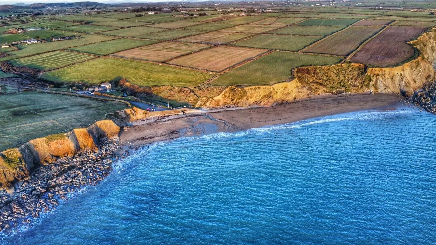 Aerial view of Kilfarrasy's coastal landscape features rugged cliffs and a sandy strand where ocean waves gently meet the shoreline. Lush green and brown fields stretch across the top, with scattered buildings near the cliffs.