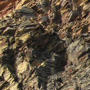 Close-up of layered, textured rock formations at Kilfarrasy Strand, Ireland, featuring various shades of brown, gray, and hints of yellow. The surface appears rugged, showcasing different stratifications and natural weathering patterns.