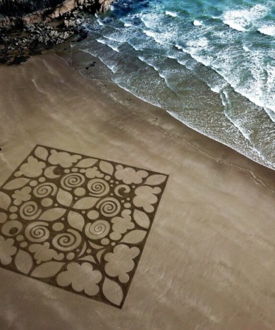 Aerial view of a beach on the Copper Coast, featuring intricate sand art with spirals and floral patterns inside a square. The artwork is near the shoreline, with waves approaching and rocks nearby, inviting onlookers to make sand art in this inspiring setting.