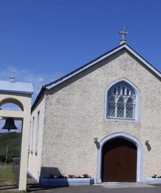 St. Mary's Church in Bonmahon is a charming beige sanctuary with a brown door and blue trim, featuring arched windows and a cross on the roof. A historic bell hangs nearby under the clear blue sky, complemented by vibrant flower beds at the entrance.