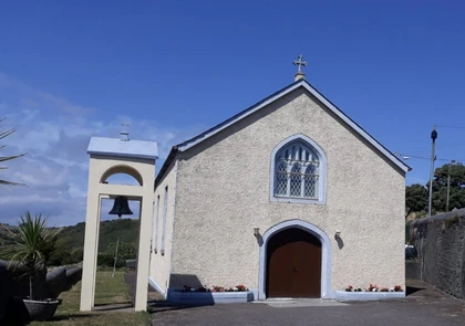 A quaint, light-colored chapel, reminiscent of St. Mary’s Church in Bonmahon's history, stands under a clear blue sky. It features an arched entrance and a stained-glass window. A small bell tower is visible to the left, surrounded by a grassy area and a few trees.