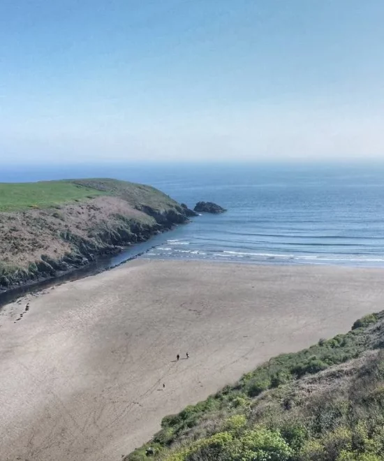 An aerial view of Stradbally Cove reveals a coastal landscape with a sandy beach edged by grassy cliffs. A river gracefully curves into the sea, while two small figures stroll along the shore under a clear blue sky.