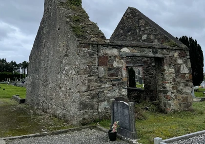 A weathered stone ruin of an ancient church stands in a cemetery in the Village of Kill, Ireland. The structure has a partially collapsed roof and walls with a doorway and an open window. Gravestones are visible nearby, and the sky is overcast.