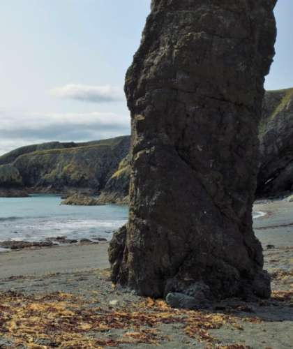 A tall rock formation stands on a sandy beach at Tra na mBo, with the ocean and rocky cliffs in the background. Seaweed is scattered across the sand, and the sky is partly cloudy.