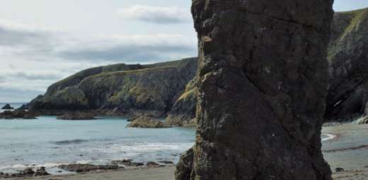 A rocky coastal scene features the majestic Tra na mBo rock formation in the foreground. In the background, imposing cliffs overlook a calm sea under a cloudy sky, while the shore is adorned with smaller rocks and pebbles.