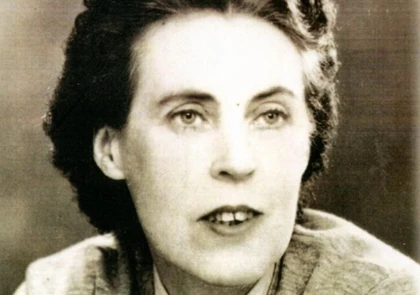 Black and white portrait of a woman with short, wavy dark hair, wearing a light-colored top. Her expression is neutral as she gazes slightly away from the camera, reminiscent of Una Troy, the acclaimed author.