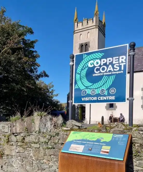 A stone building with a clock tower stands in the background under a clear blue sky. In the foreground, a large sign reads "Copper Coast UNESCO Global Geopark Visitor Centre," highlighting its role in management. A smaller informational board is mounted on a stone wall.