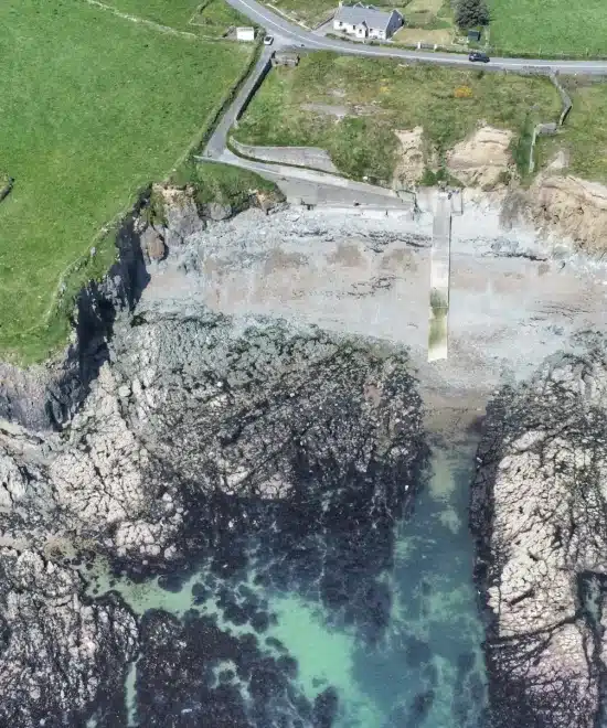 Aerial view of a coastal landscape reveals fascinating geology with rocky cliffs and a shoreline at low tide. A narrow pier extends into the water, surrounded by grass-covered land. At the top, a small road and buildings dot the scene. The clear water showcases visible seaweed formations.